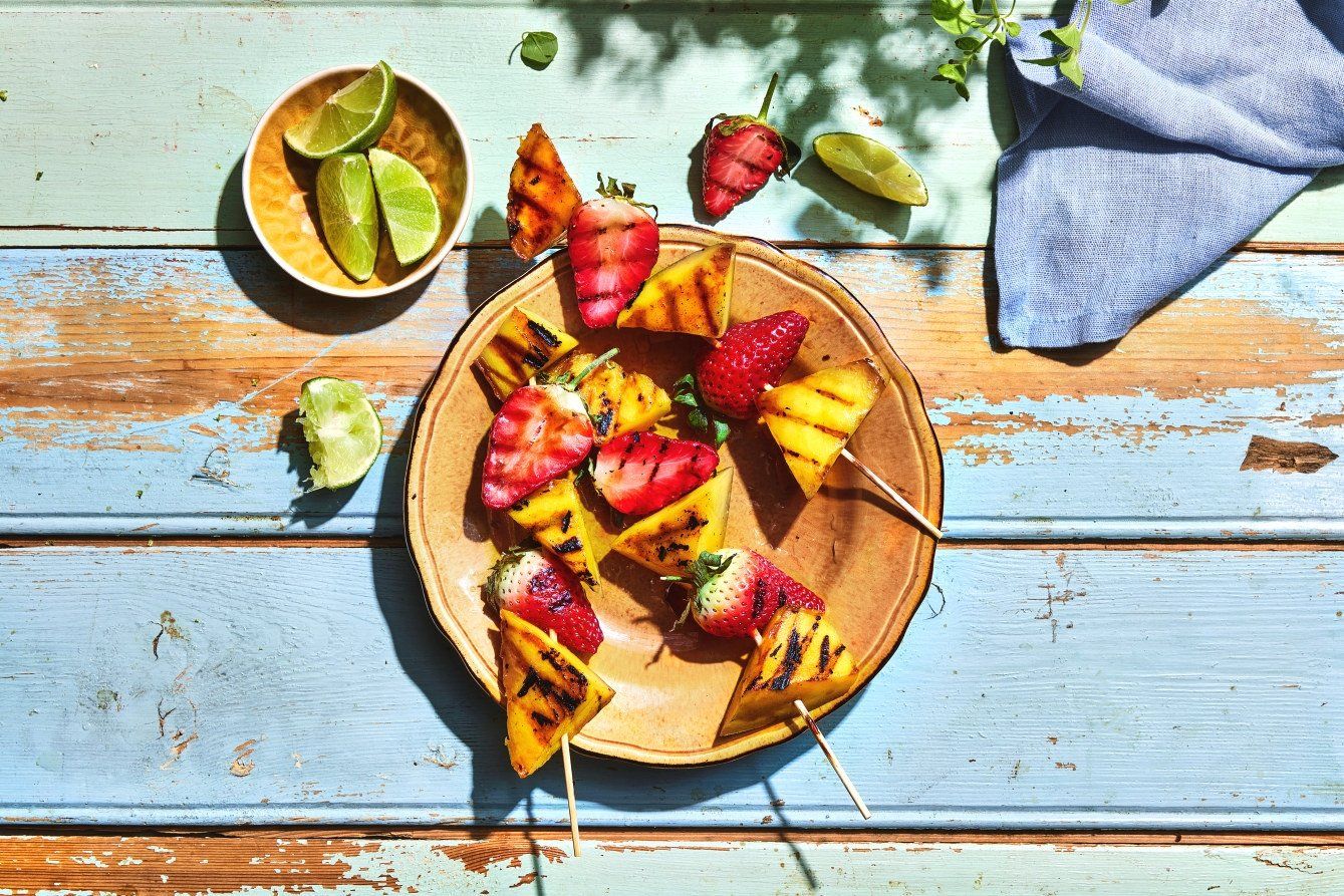 Fargerike grillspyd med biter av ananas og hele jordbær, merket med tydelige grillstriper. Spydene ligger på en brun tallerken plassert på et rustikt, lyseblått trebord.