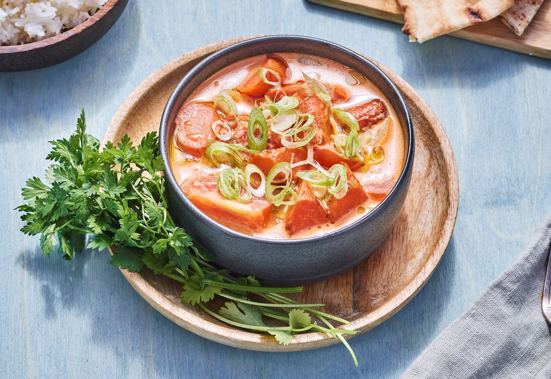 En kremet tomatsuppe servert i en mørk bolle, toppet med vårløk og tomatbiter, med frisk persille ved siden av på et rundt trefat.