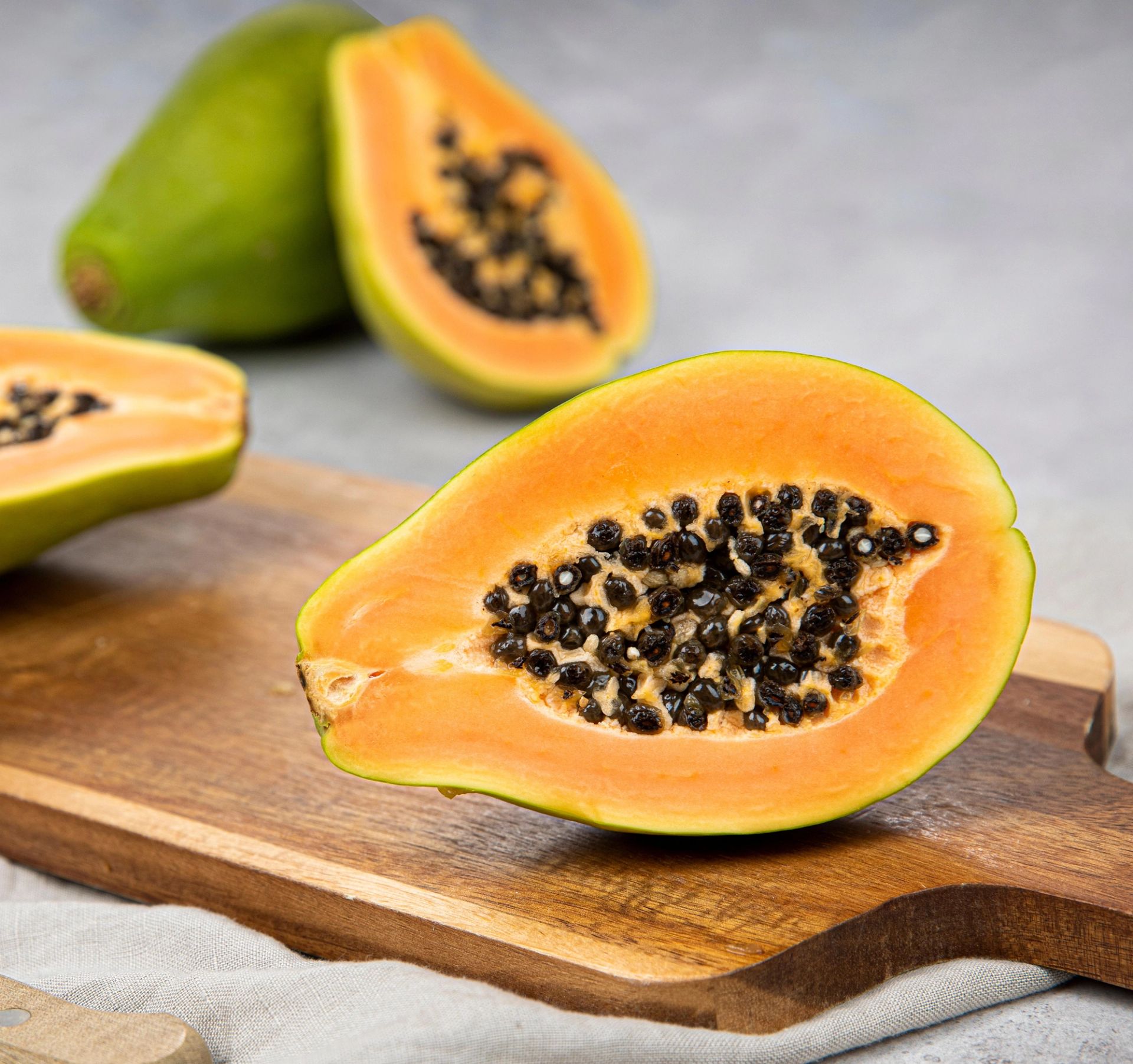 Halv papaya med oransje fruktkjøtt og svarte frø ligger på et trefjøl, med flere hele og delte papayaer i bakgrunnen på en lys overflate.