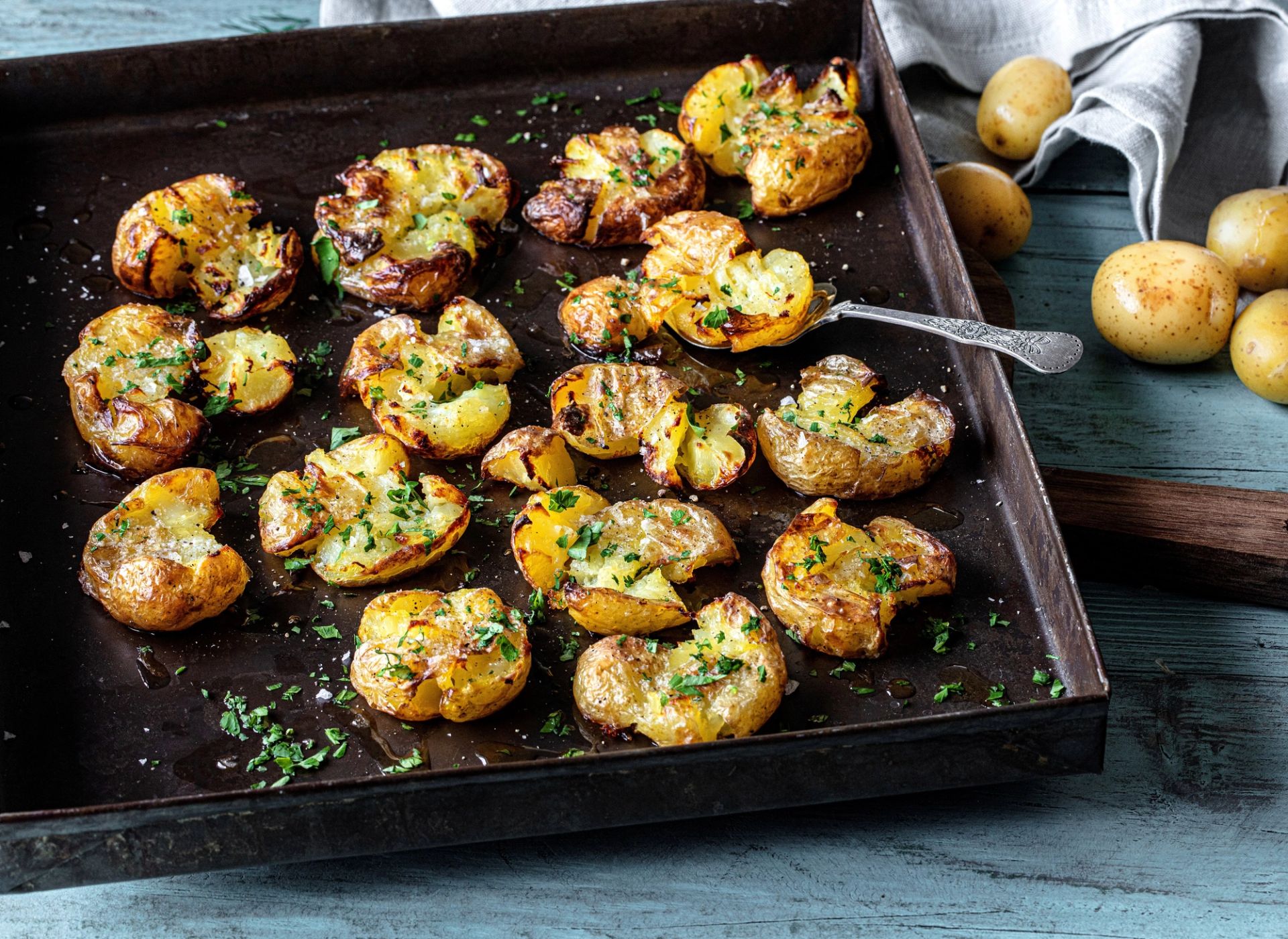 Knuste, ovnsbakte poteter ("smashed potatoes") på et mørkt stekebrett av metall. Potetene er gylne og sprø med dryss av havsalt og finhakket persille.