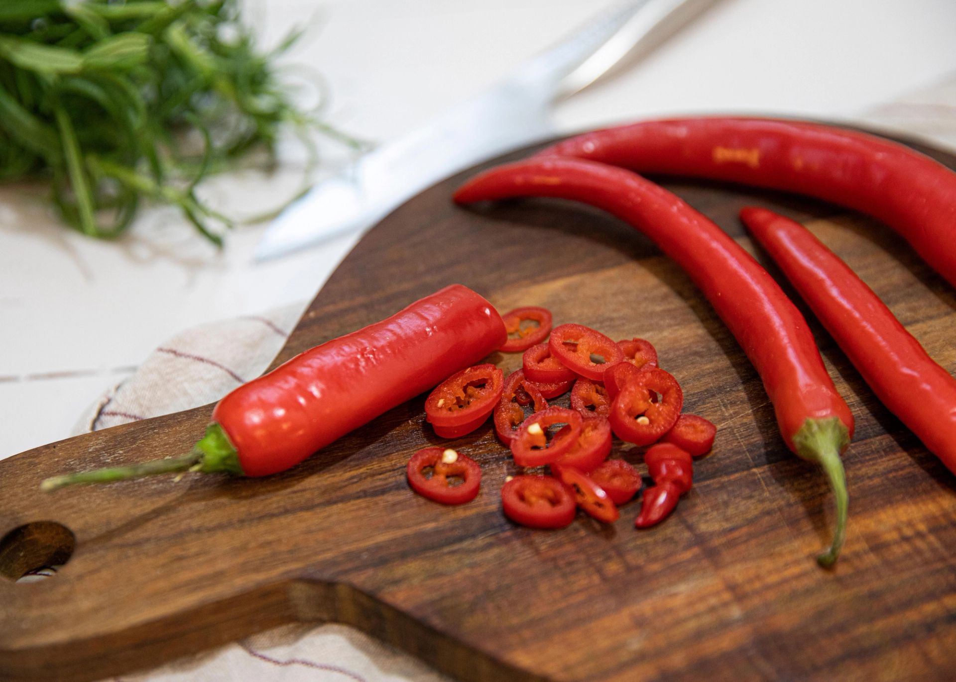 Rød chilipepper og skiver av chili ligger på et rundt skjærebrett i tre, med en kniv og grønne urter i bakgrunnen på en lys kjøkkenbenk