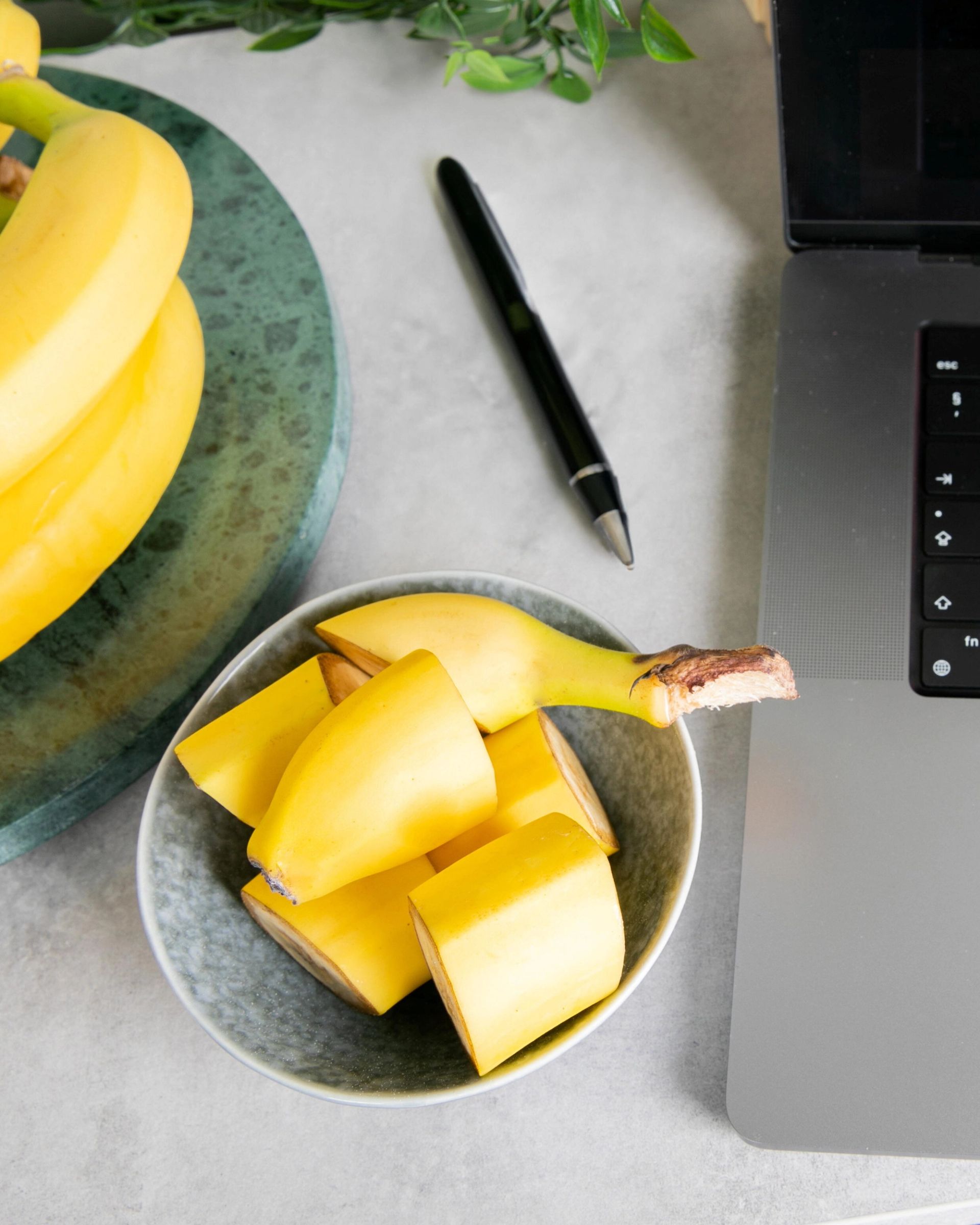 Skål med oppskårne bananbiter står ved siden av en bærbar PC på et skrivebord. En penn ligger ved siden av skålen, og flere hele bananer er synlige i bakgrunnen, noe som gir inntrykk av et enkelt og praktisk mellommåltid under arbeid eller studier.