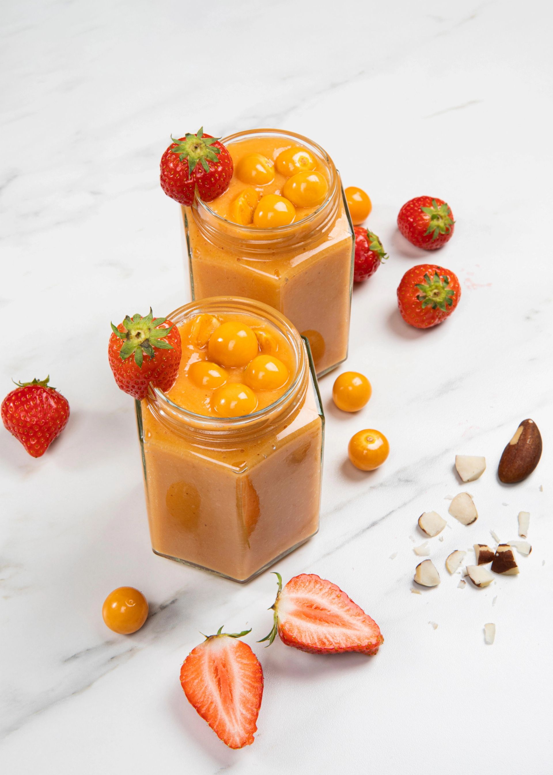 To glass med oransje smoothie laget av physalis og jordbær, toppet med hele physalisbær. Hvert glass har et jordbær på kanten. Rundt glassene ligger hele og halve jordbær, physalisbær og noen hakkede nøtter på en hvit marmorlignende overflate.