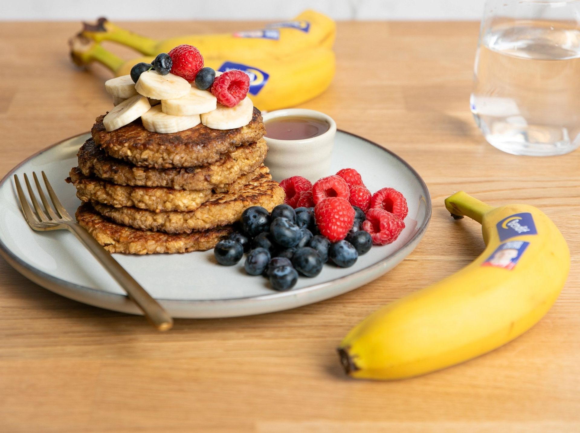 Stabel med havre- og bananpannekaker toppet med bananskiver, bringebær og blåbær, servert med sirup, friske bær og bananer på et bord.
