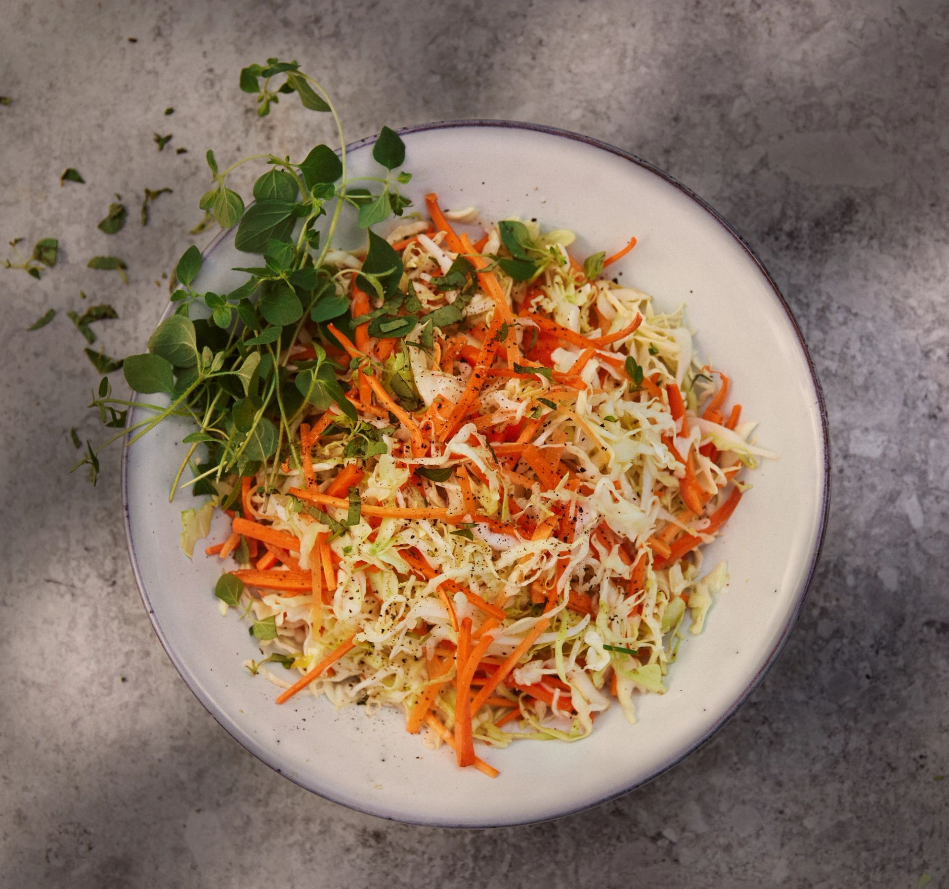 En fargerik råkostsalat servert i en rund, lys grå tallerken, fotografert ovenfra. Salaten består av finsnittet hvitkål og tynne strimler av oransje gulrot, lett krydret med kvernet svart pepper. En stor kvast med frisk, grønn oregano ligger på venstre side av tallerkenen, og noen løse blader er spredt på den mørkegrå, marmorerte bakgrunnen.