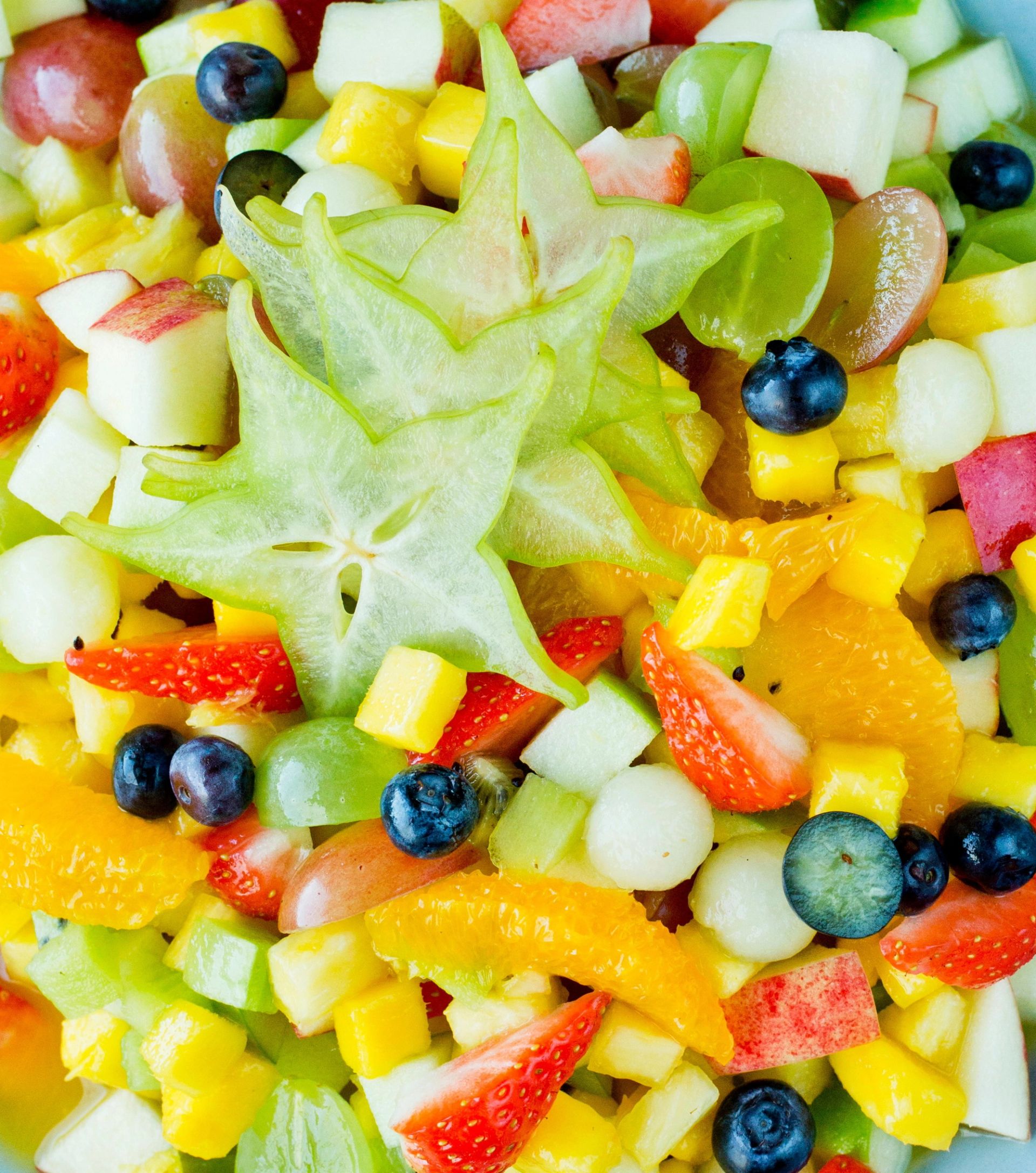 Nærbilde av fargerik fruktsalat med terninger av melon, mango og eple, skiver av jordbær, blåbær og en stjerneformet skive av stjernefrukt på toppen.