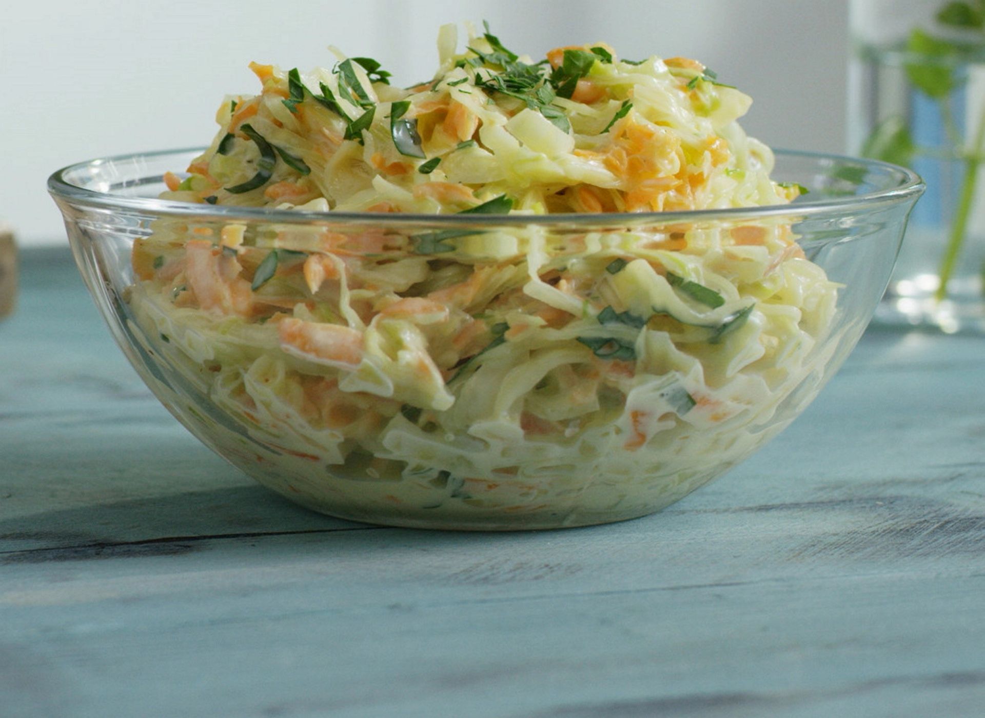 Kremet coleslaw med kål, gulrot og persille i en glasskål, servert på et turkist trebord.