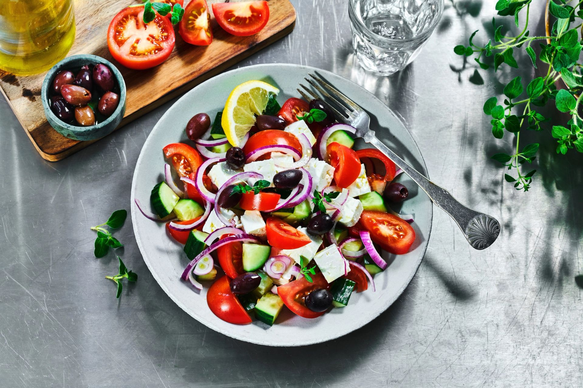 Tallerken med gresk salat med tomater, agurk, rødløk, svarte oliven og fetaost, pyntet med sitronskive. Tallerkenen står på et metallbord, med en gaffel ved siden av. I bakgrunnen ligger et skjærebrett med tomater, en skål med oliven, en flaske olivenolje, et glass vann og friske urter.