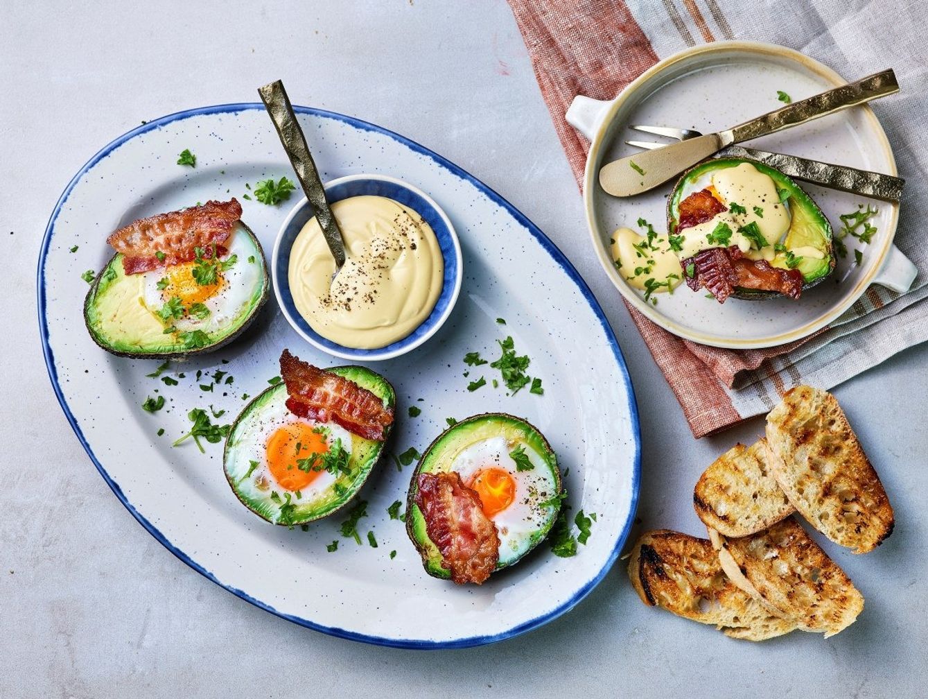 Avokadohalvdeler bakt med egg og toppet med sprøstekt bacon, servert med majones og grillet brød på siden, garnert med frisk persille på et ovalt fat og en mindre tallerken.