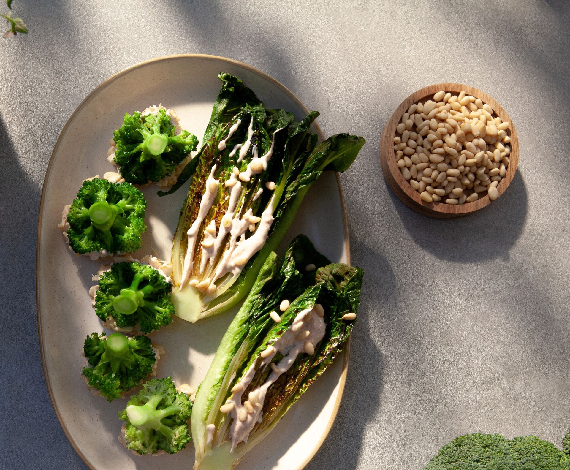 Oval tallerken med grillet hjertesalat, toppet med kremet dressing og pinjekjerner, samt små forretter med brokkolibuketter på toppen. En skål med pinjekjerner og fersk brokkoli står ved siden av.