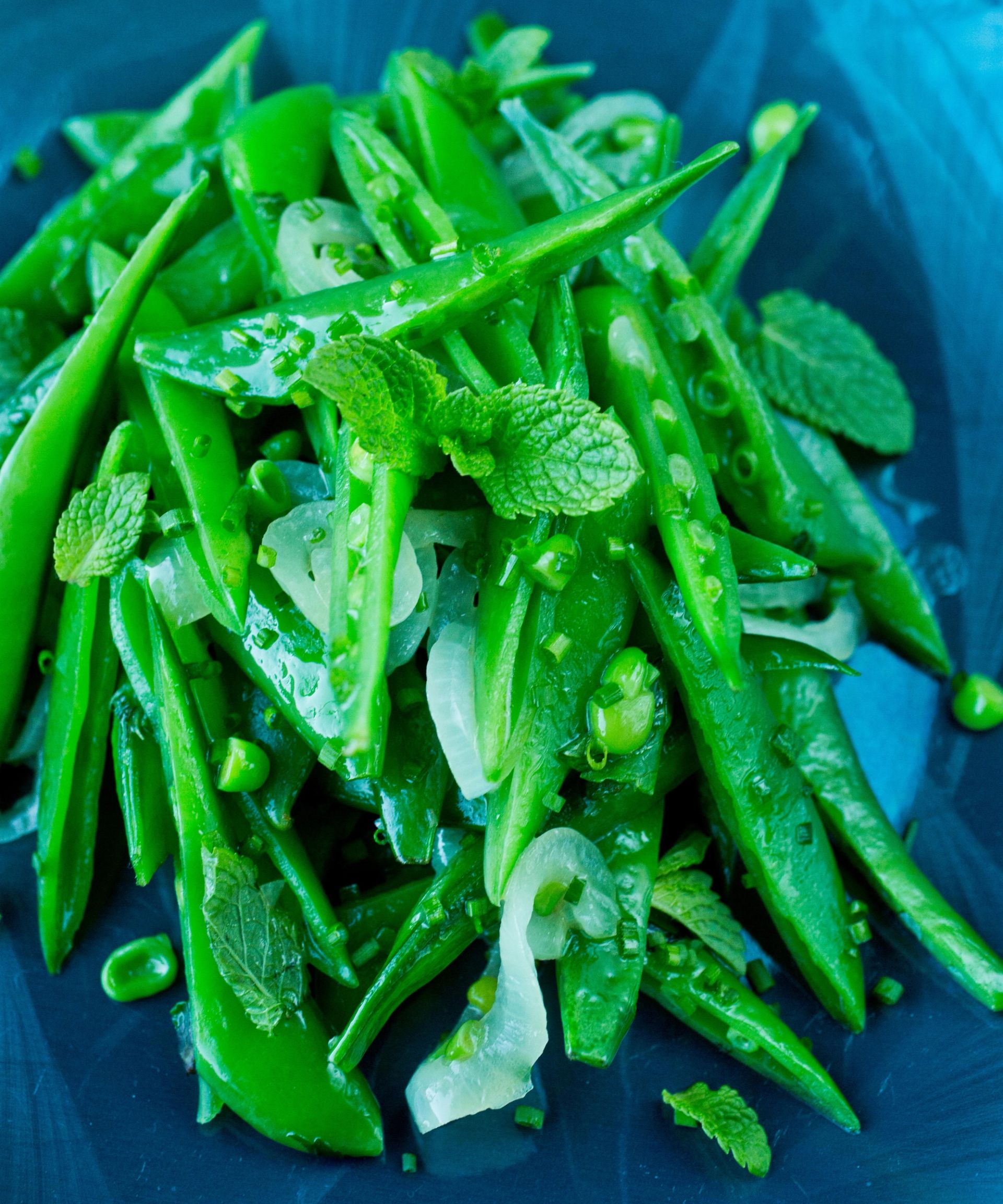 Frisk salat av grønne sukkererter og bønner blandet med gressløk og mynteblader, servert på et mørkt fat.
