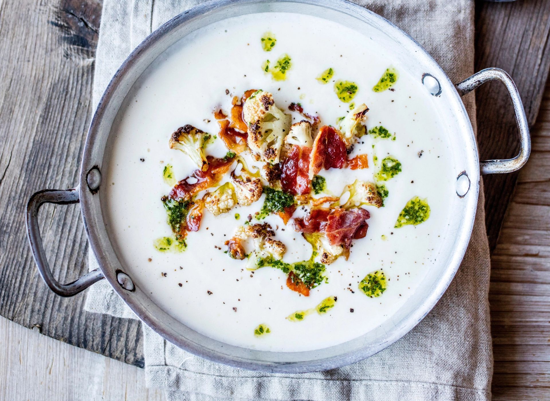 Kremet blomkålsuppe med sprøstekt bacon, ristede blomkålbuketter og grønnkålpesto servert i en rustikk metallgryte. Gryten står på et lyst linhåndkle på et værbitt trebord.