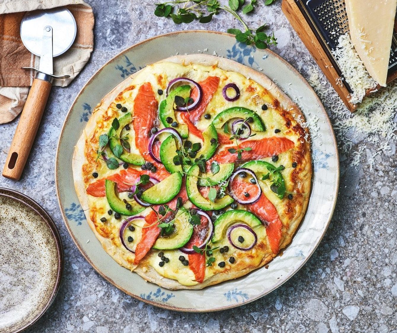 Stekt pizza toppet med røkelaks, skiver av avokado, rødløk og friske urter, servert på en tallerken med kjøkkenutstyr og revet ost rundt.