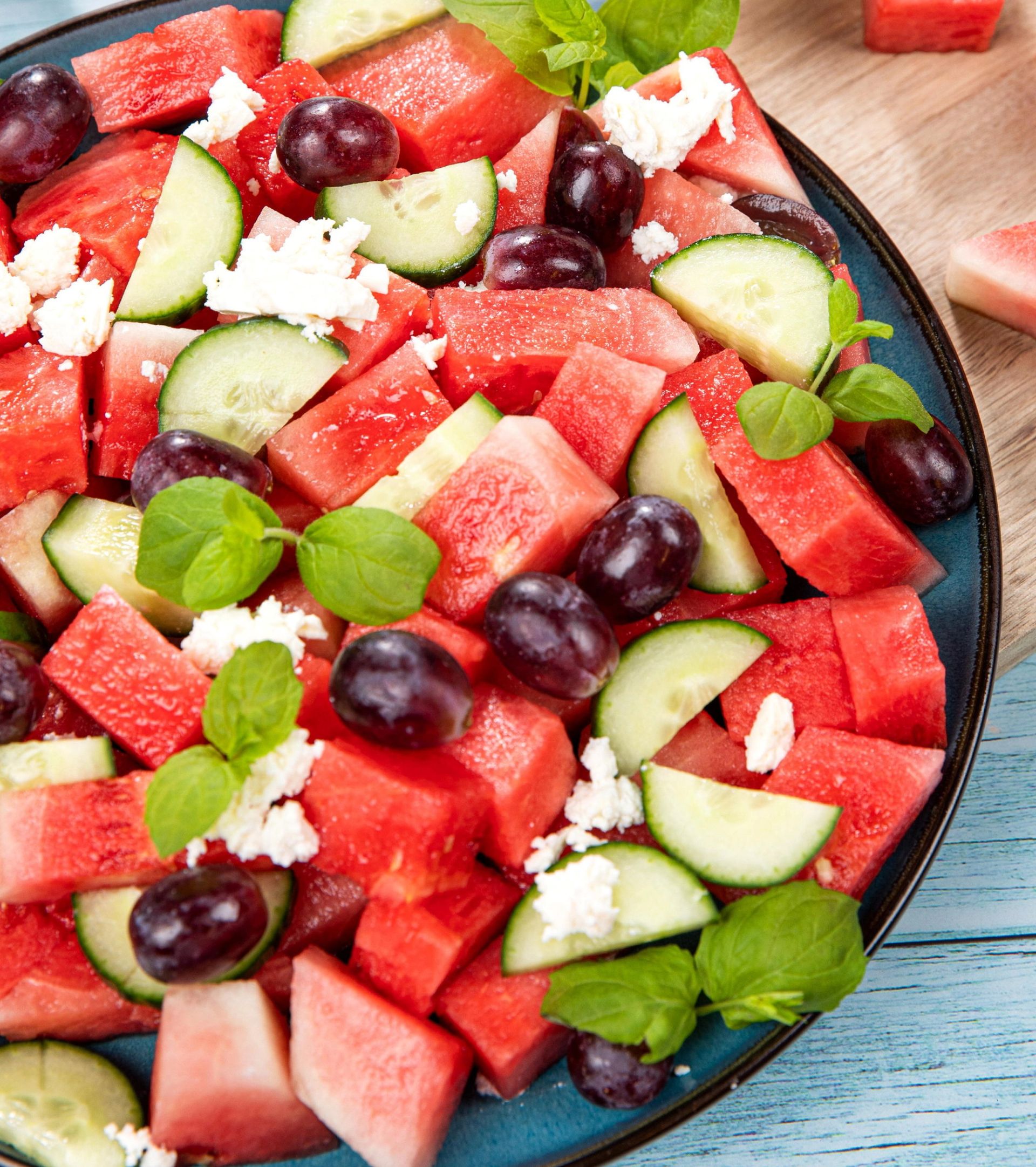 Fargerik salat med terninger av vannmelon, skiver av agurk, mørke druer, frisk mynte og smuldret hvit ost, servert i en mørk skål på et lyst trebord.