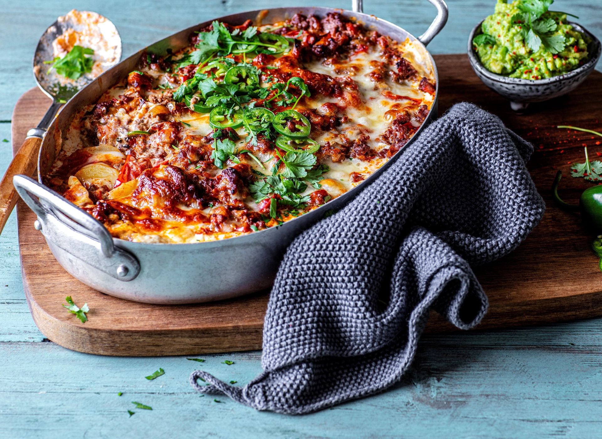 En dampende potet- og tacograteng i en oval metallform, fylt med kjøttdeig, ost og potetskiver. Gratengen er toppet med frisk koriander og skiver av grønn chili. Formen står på et trebrett med et grått kjøkkenhåndkle ved siden av, og i bakgrunnen står en liten skål med guacamole.