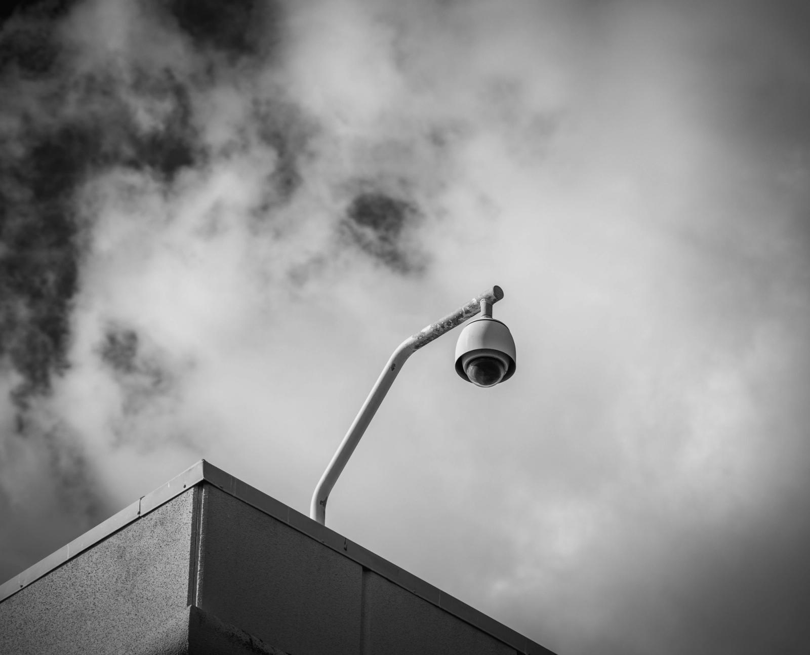 A single CCTV camera on the corner of a retail building in black and white.