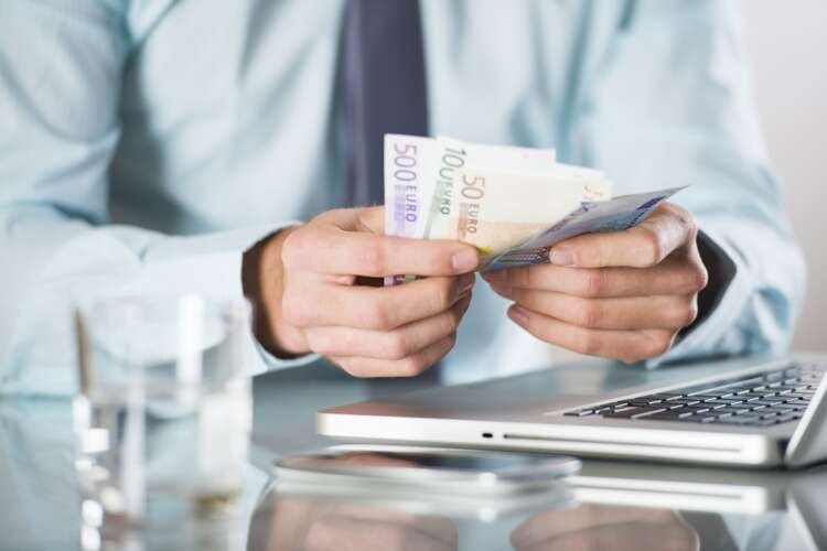Businessman using laptop with money in his office, illustrating cross-border payments - Global Banking & Finance Review