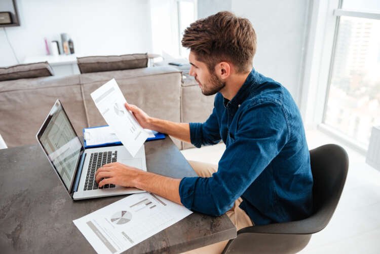 A man analyzing home finance papers while typing on a laptop - Global Banking & Finance Review