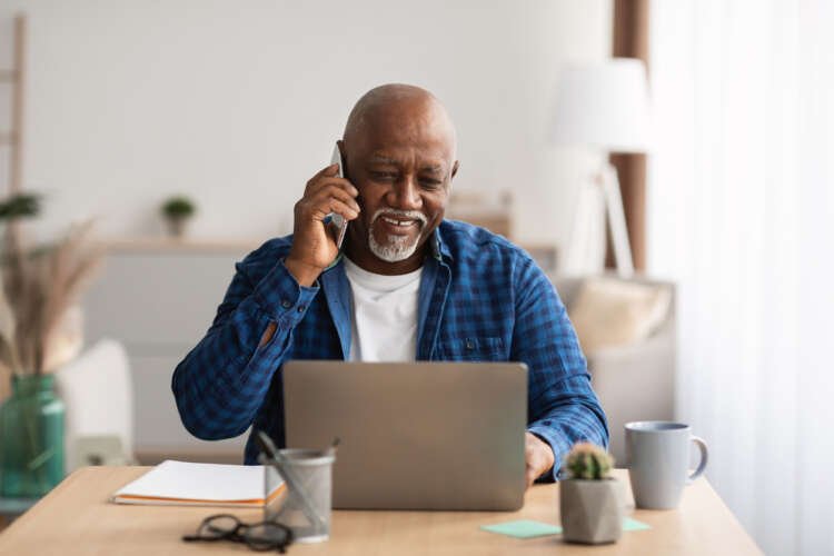 Mature African man using a laptop and cellphone, representing digital engagement in pensions - Global Banking & Finance Review