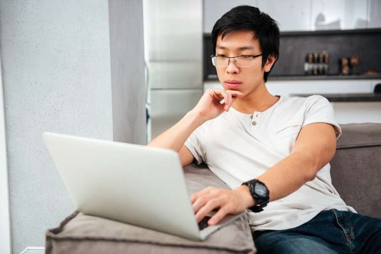 Serious Asian man with laptop on sofa analyzing money transfer trends - Global Banking & Finance Review