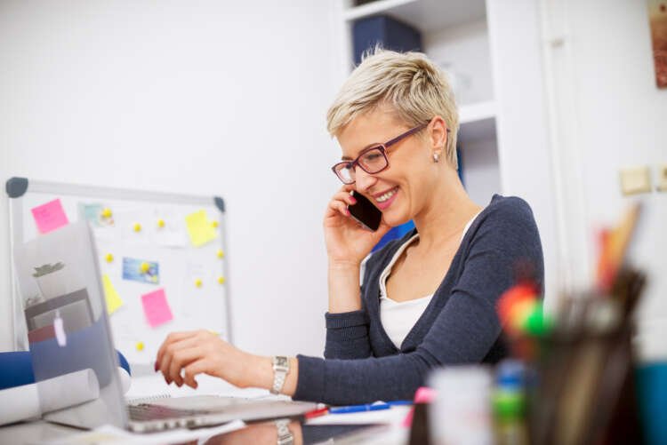 Attractive charming blonde short hair business woman talking on a mobile while sitting at office desk and working on a laptop.