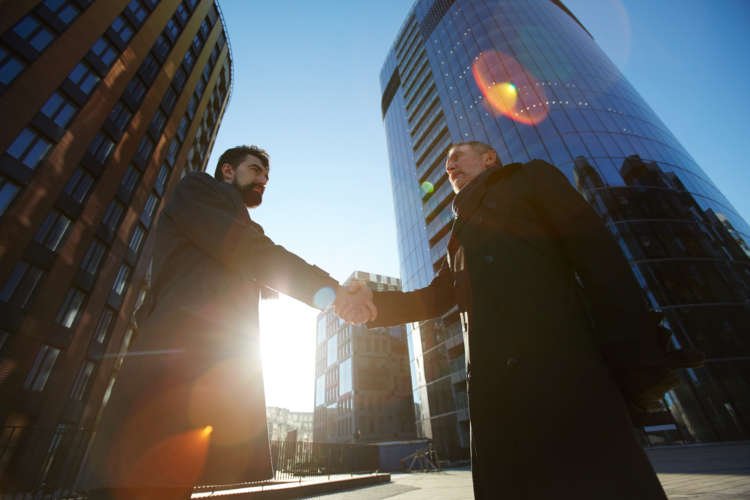 Two contemporary employers shaking hands outdoors near skyscrapers, symbolizing business partnerships - Global Banking & Finance Review