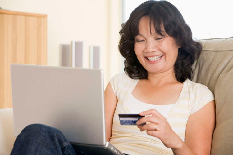 Woman smiling while using a laptop and holding a credit card at home - Global Banking & Finance Review