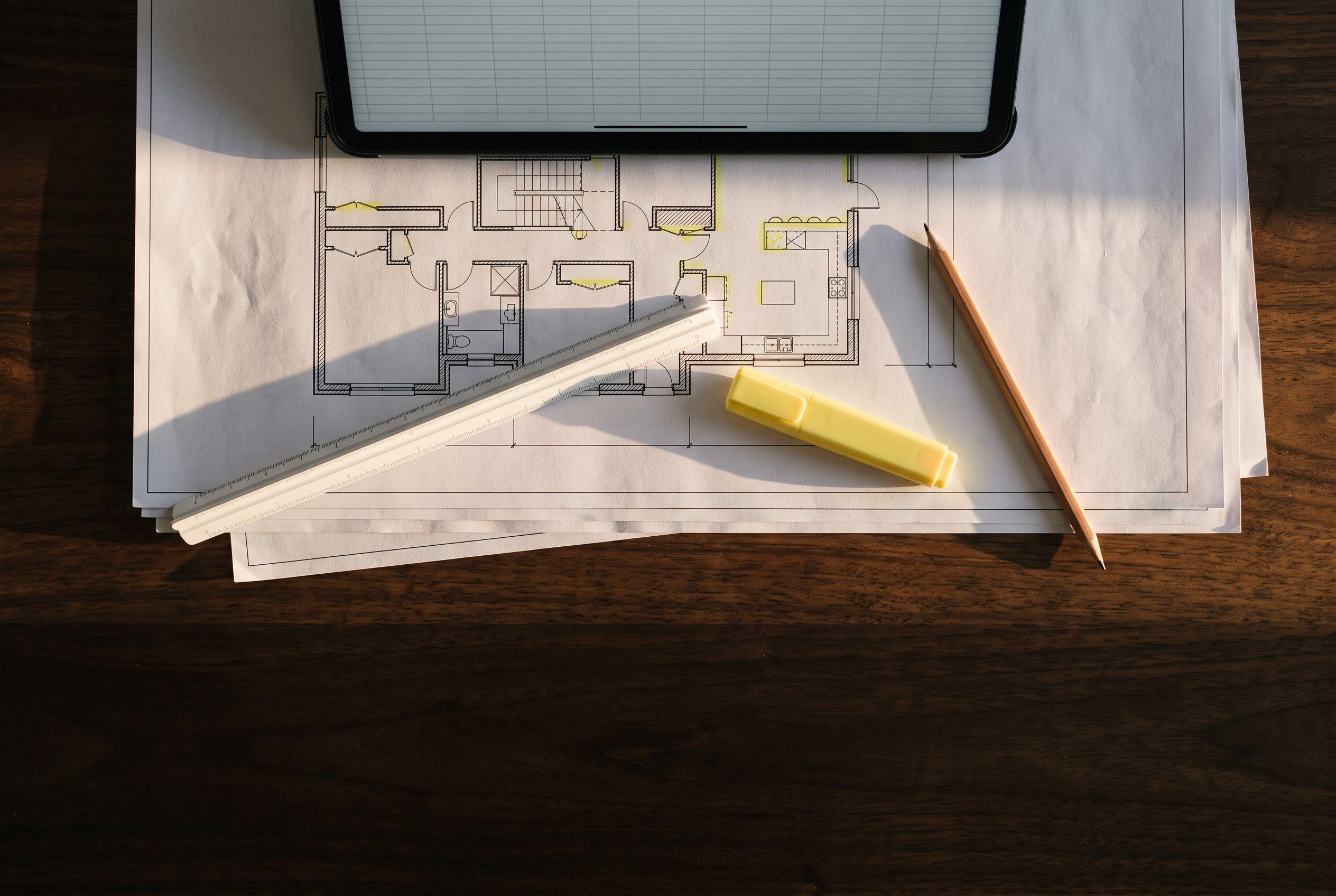 Floor plans marked up with a highlighter, beside a scale rule, a pencil and a tablet showing a spreadsheet