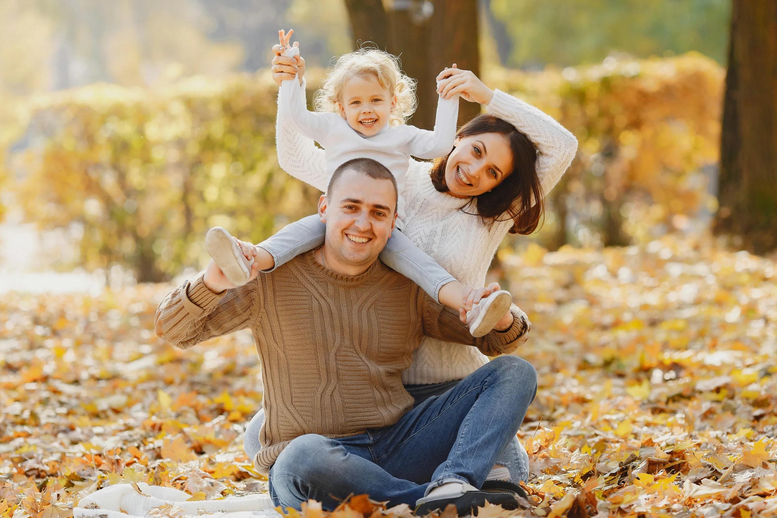 Smiling family of three with a child on a man's shoulders in an autumn park.