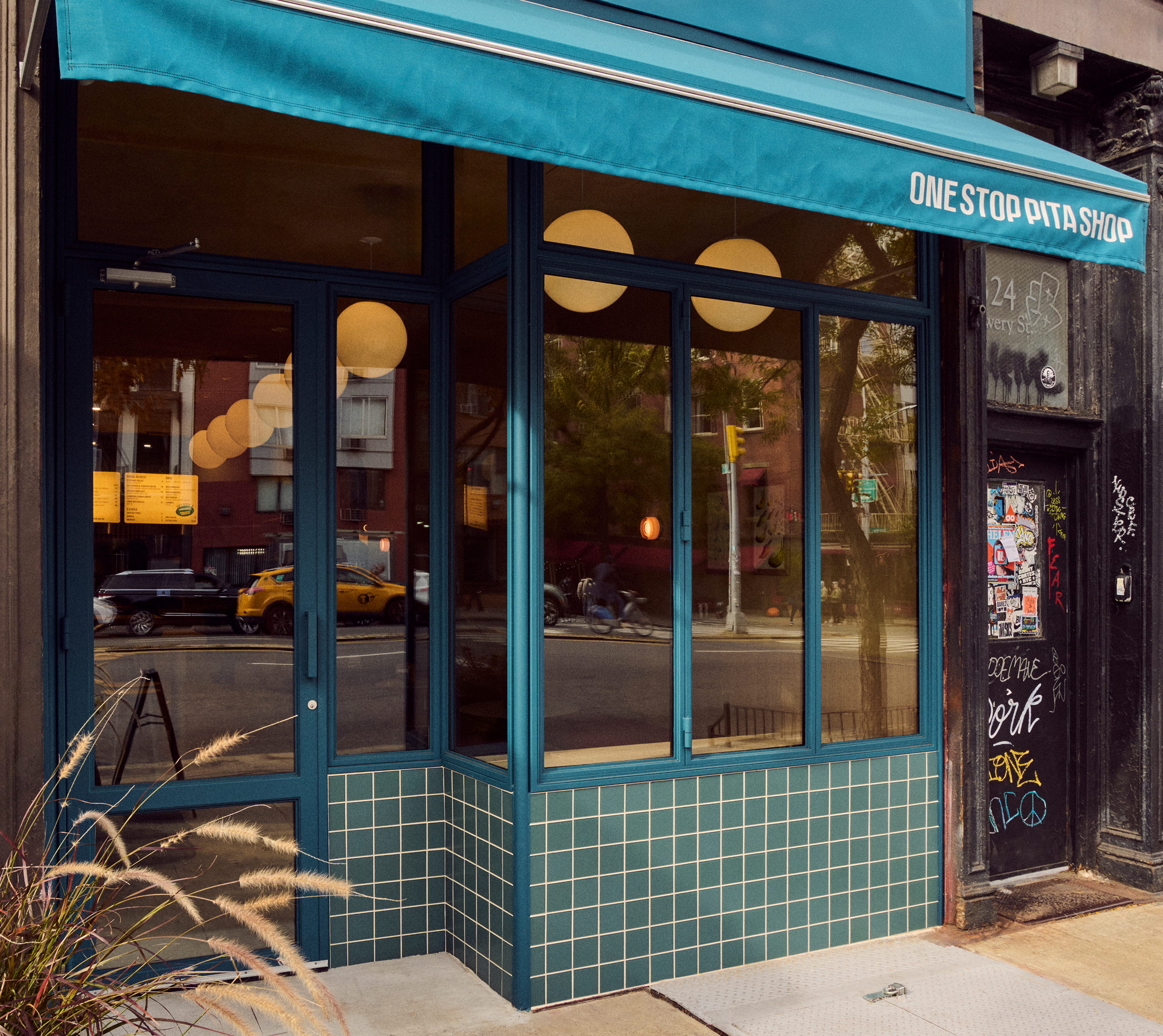 Storefront of "ONE STOP PITA SHOP" with a teal awning, windows, and tiled base, reflecting the street.