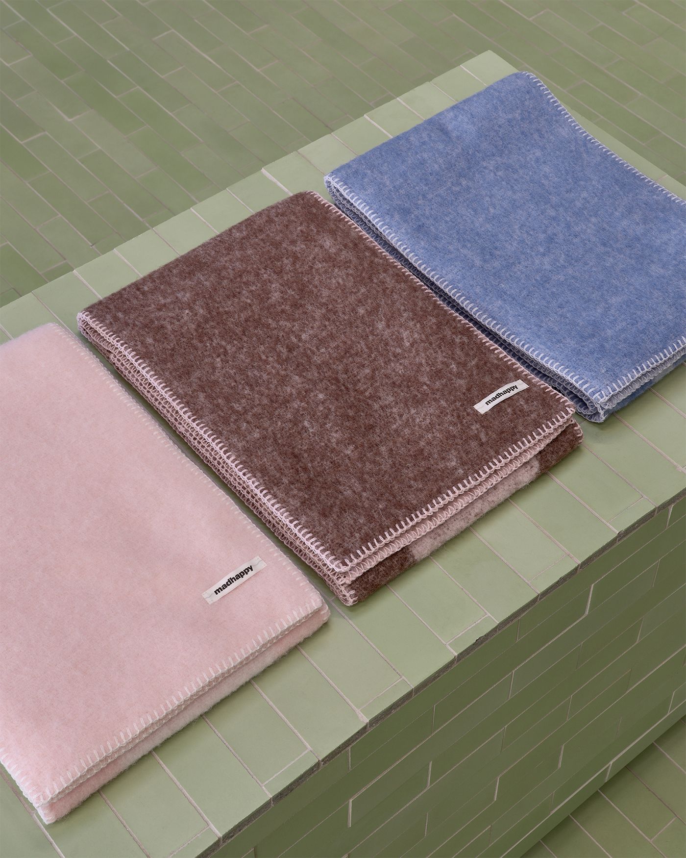 Three folded blankets in pink, brown, and blue with "madhappy" tags and white stitching on a green tiled surface.