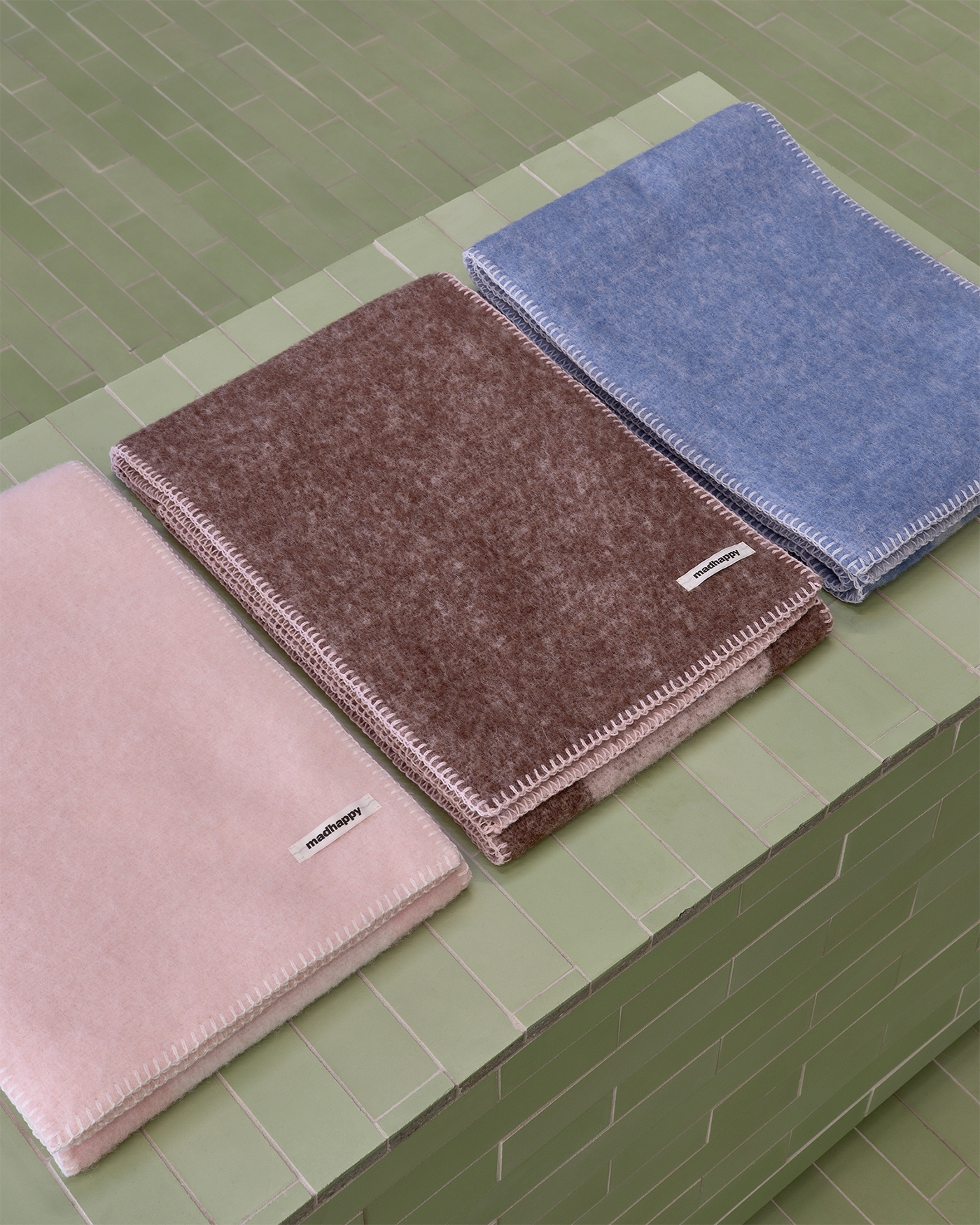 Three folded blankets in pink, brown, and blue with "madhappy" tags and white stitching on a green tiled surface.