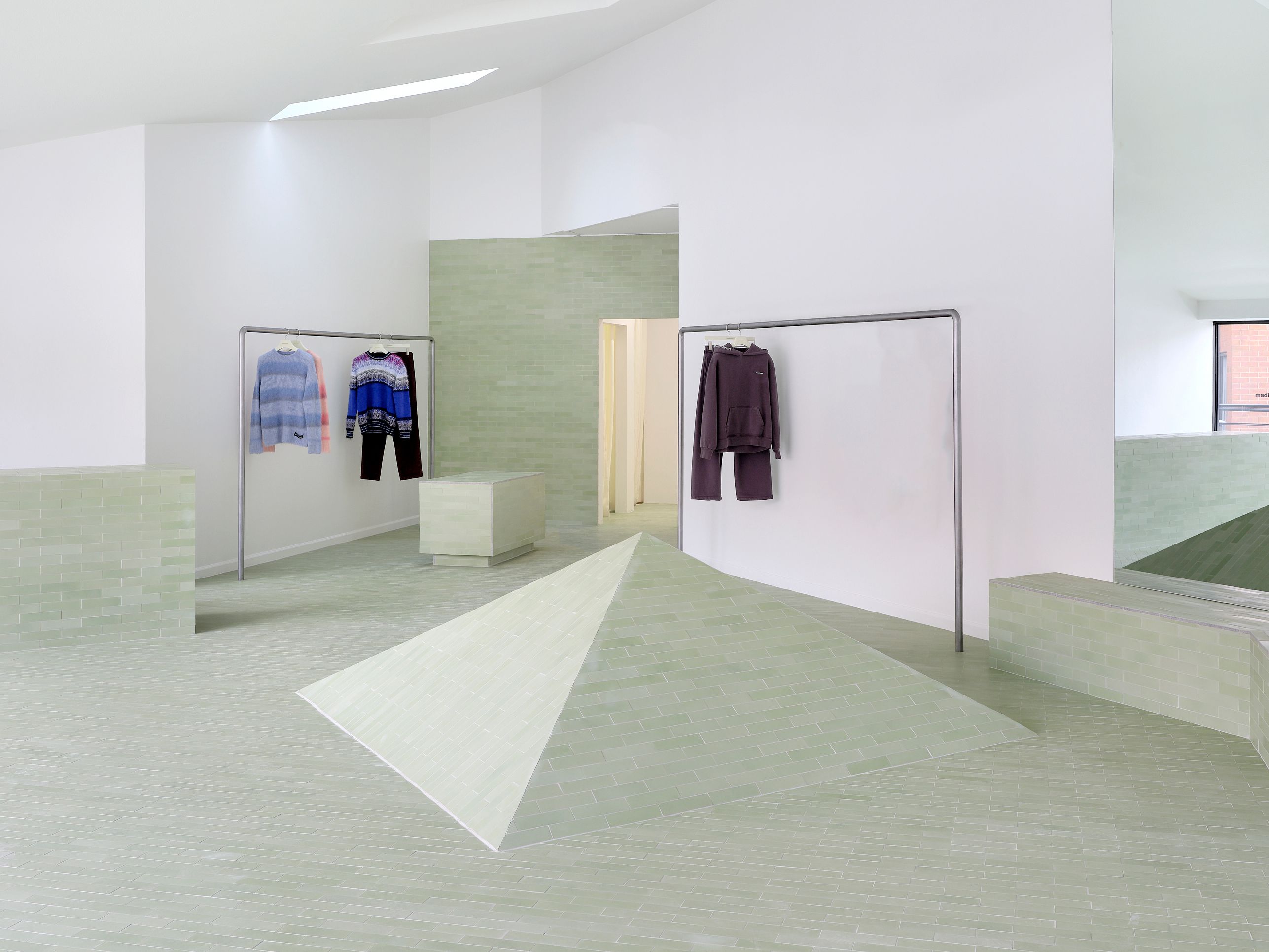 Minimalist boutique with sage green tiled floor, a central pyramid, and clothes displayed on racks.