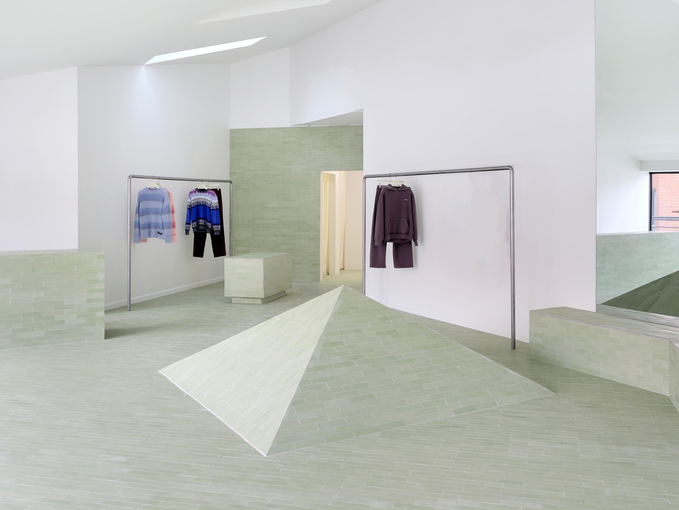 Minimalist boutique with sage green tiled floor, a central pyramid, and clothes displayed on racks.
