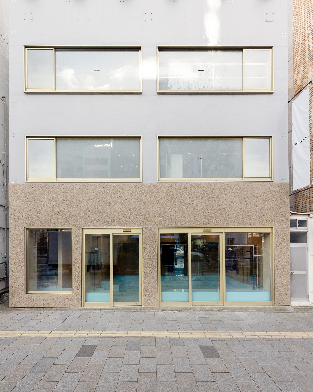 A two-story building facade with a light grey upper section and a textured brown lower section, featuring gold-framed windows and glass doors.