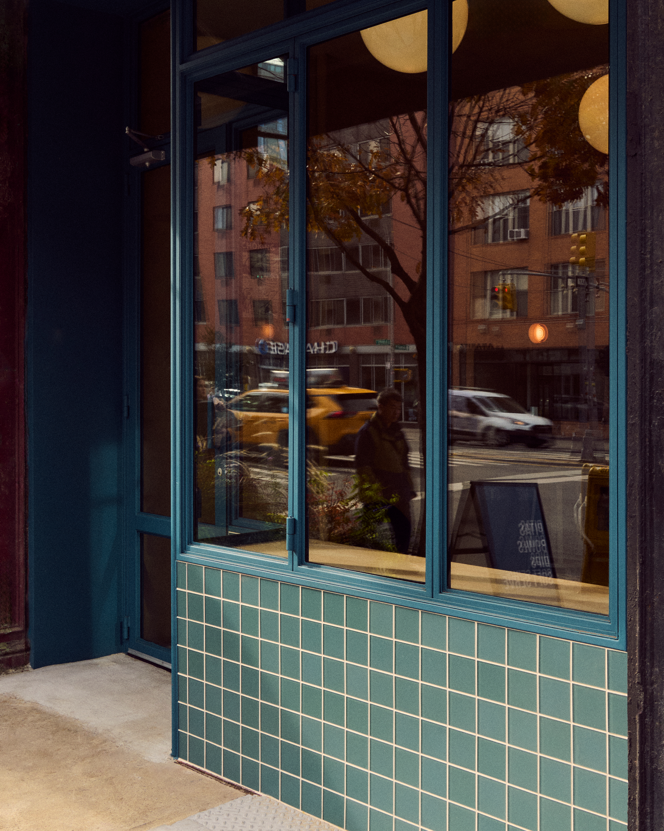 Teal-framed windows and a tiled facade reflect a city street with people, vehicles, and brick buildings.