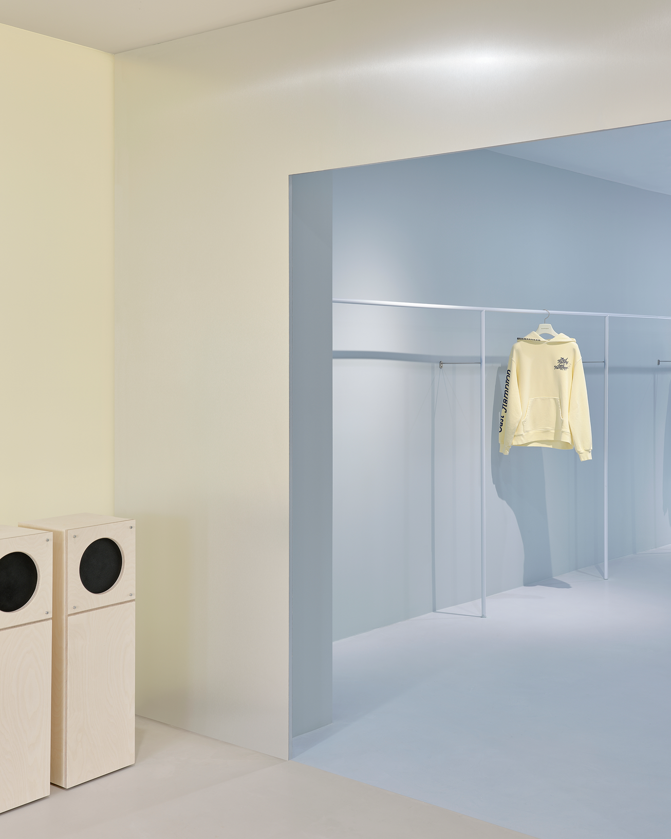 A two-tone room with a pale yellow wall and light wooden speakers on the left, and a pale blue area with a yellow hoodie on a clothes rack on the right.