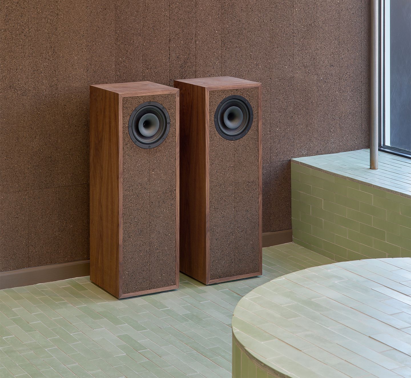 Two wood and cork floor-standing speakers against a cork wall, on a light green tiled floor with a curved tiled bench.