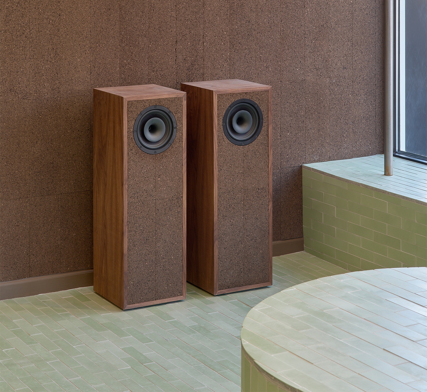 Two wood and cork floor-standing speakers against a cork wall, on a light green tiled floor with a curved tiled bench.