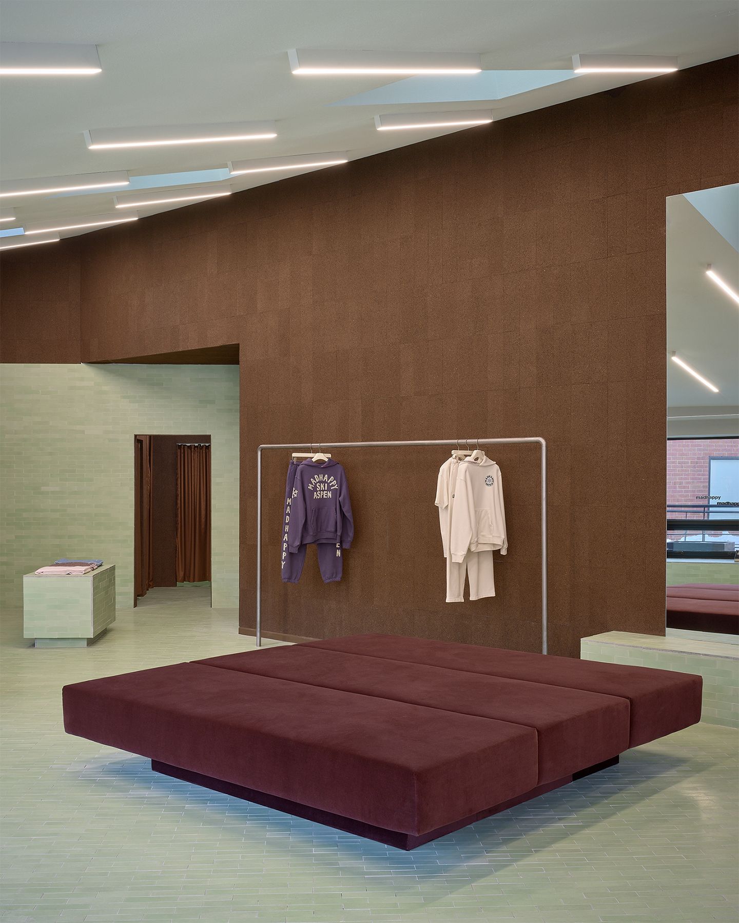Modern retail space with a large maroon bench, a clothing rack, textured brown cork walls, and a light green tiled floor.