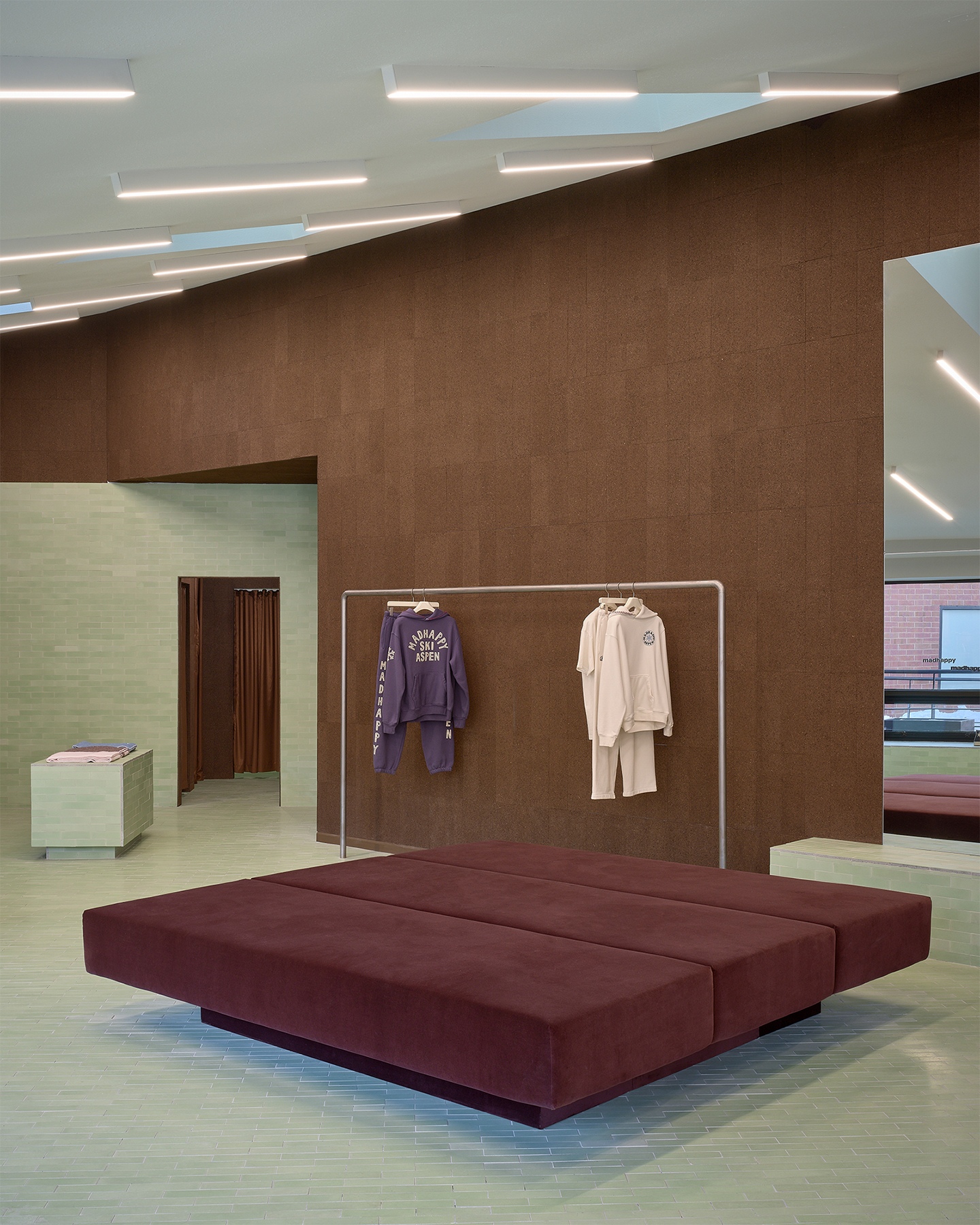 Modern retail space with a large maroon bench, a clothing rack, textured brown cork walls, and a light green tiled floor.