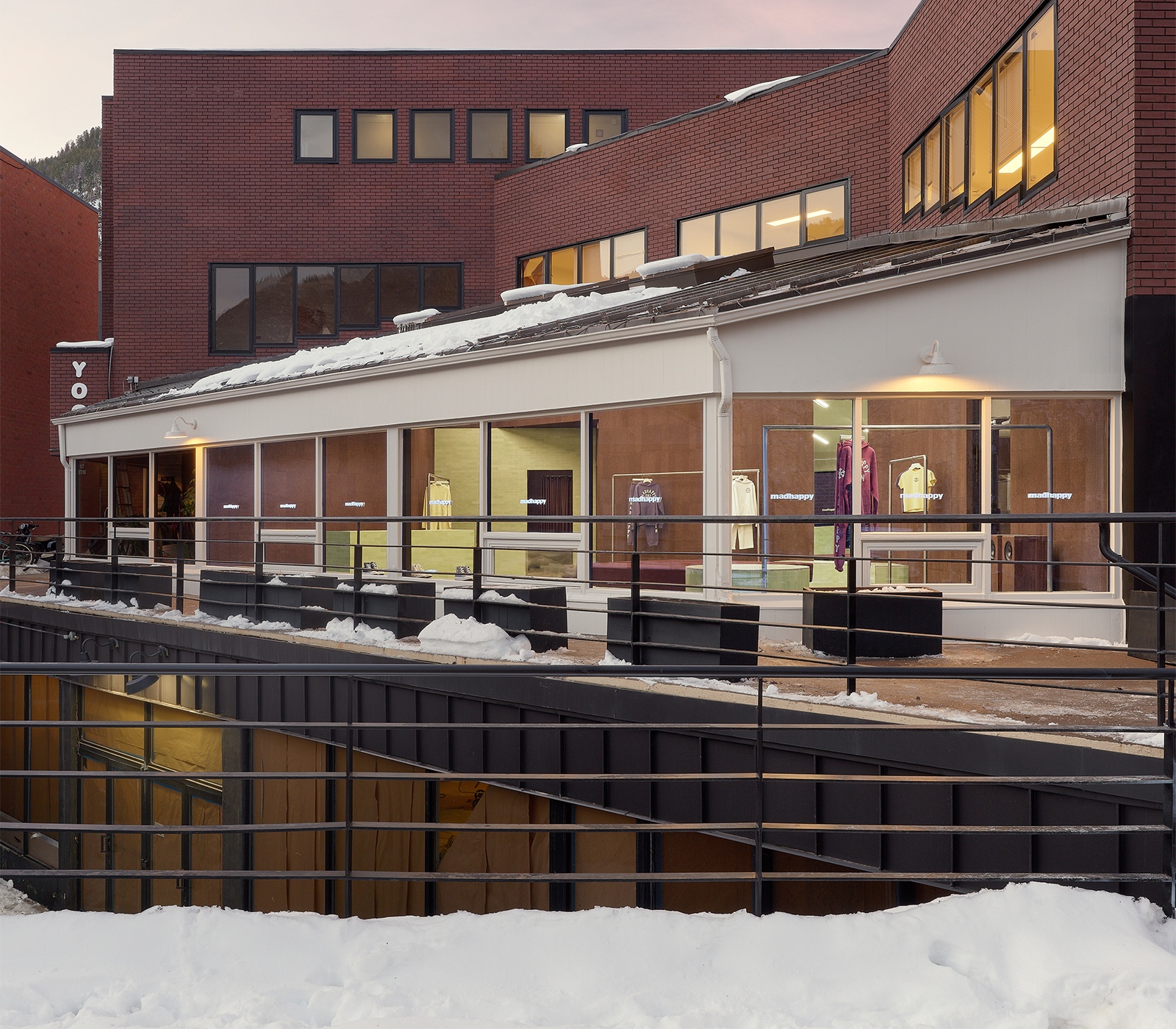 A white, glass-fronted boutique displaying clothes, with snow on the ground and a large brick building behind it. Madhappy Clothing brand store.