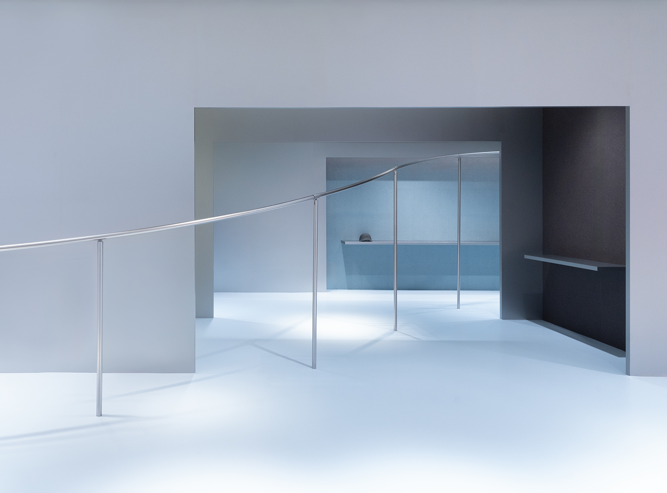 A minimalist interior with a curving silver rail, light grey walls, and blue-toned openings to other rooms with shelves.