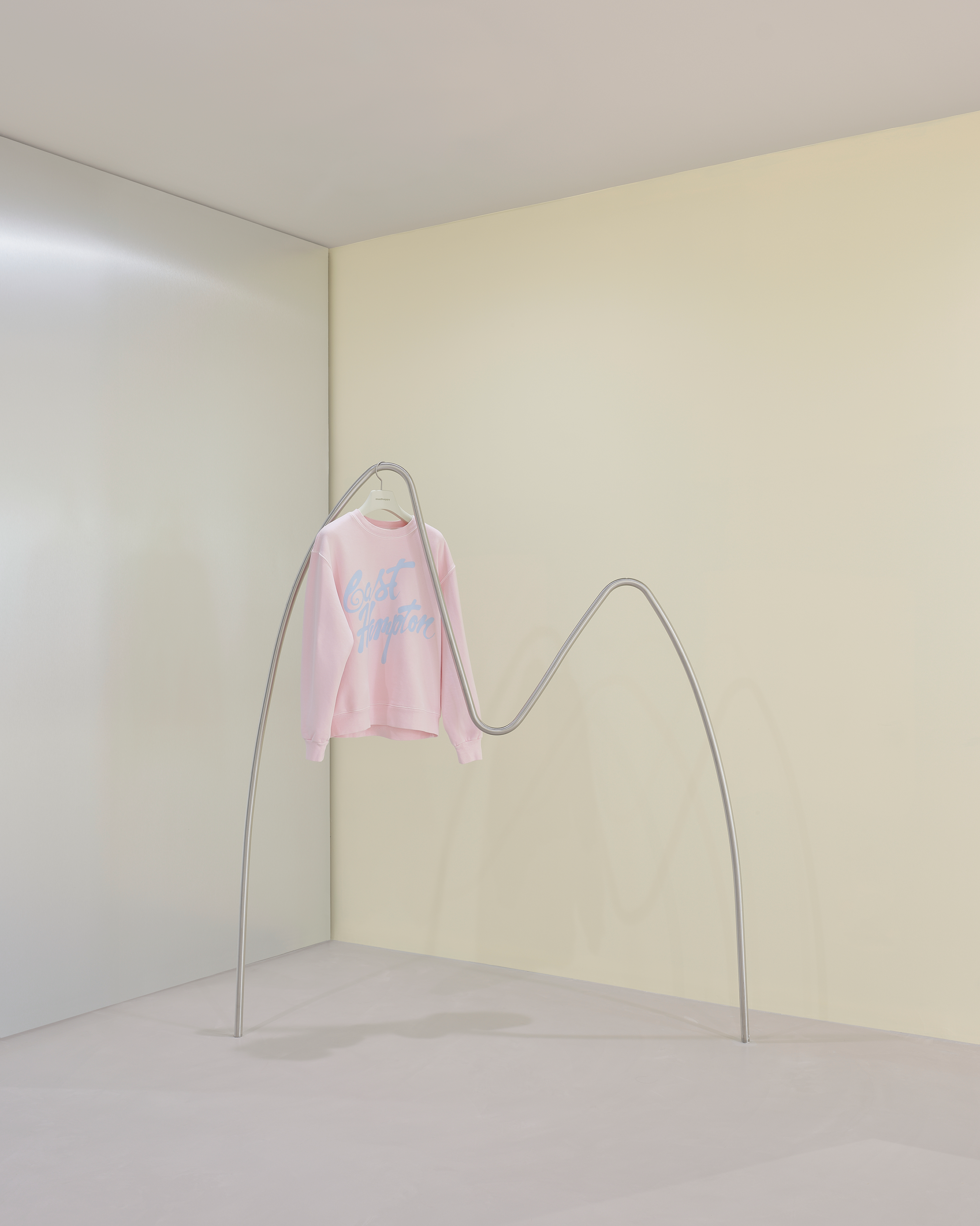 A light pink sweatshirt with blue text hangs from a sculptural, wavy silver metal clothing rack in a minimalist room.