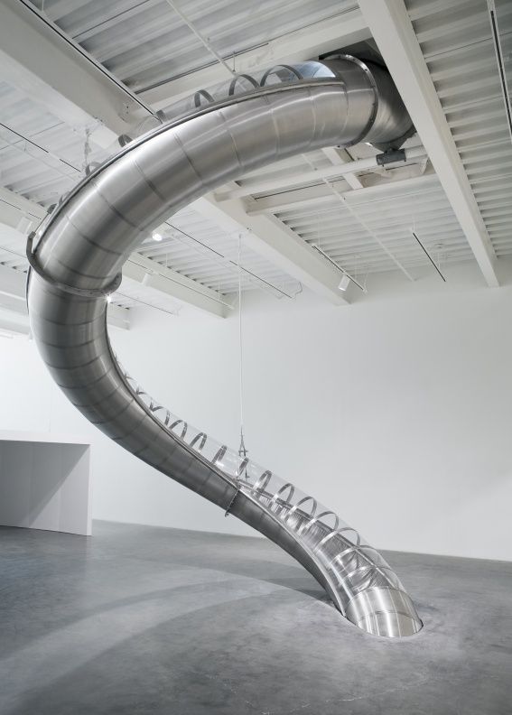 A large, curved silver slide with transparent sections descends from a high white ceiling, embedding into a concrete floor.