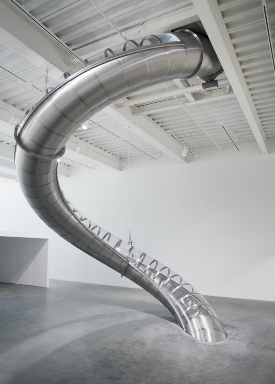 A large, curved silver slide with transparent sections descends from a high white ceiling, embedding into a concrete floor.