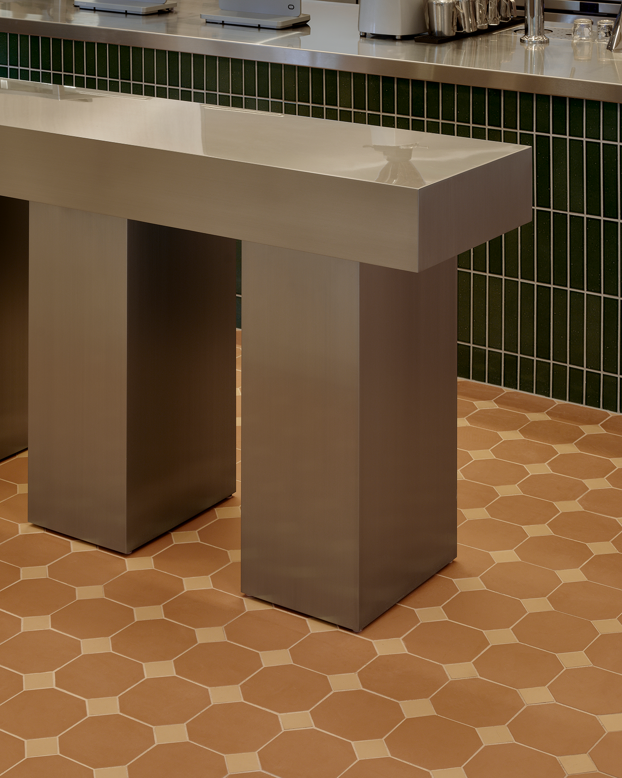 Modern interior with a sleek metal counter, dark green subway tiles, and an octagonal terracotta floor.