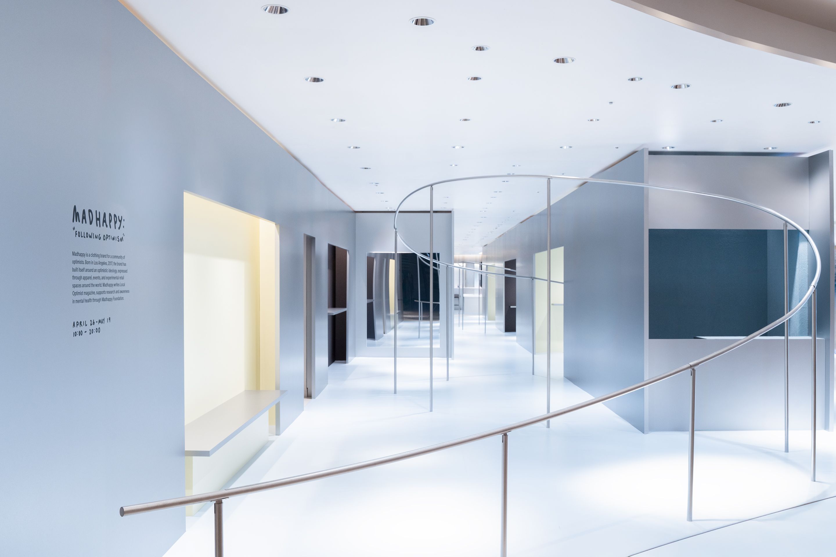 A sleek, modern interior with light gray walls, white floors, a curved metal railing, and "MADHAPPY" text on the left wall.