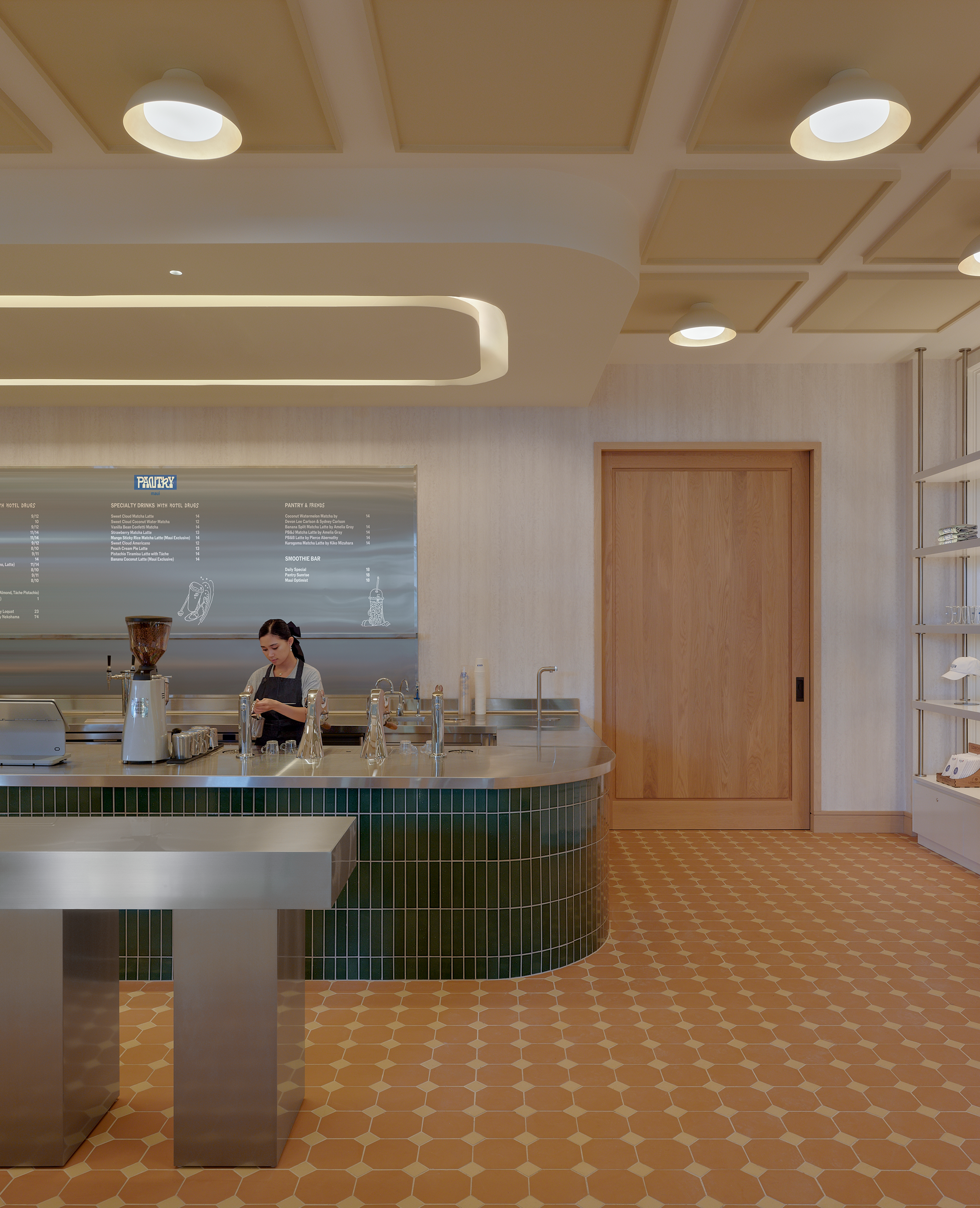 A barista works behind a curved green-tiled counter in a modern cafe with patterned terracotta floors.