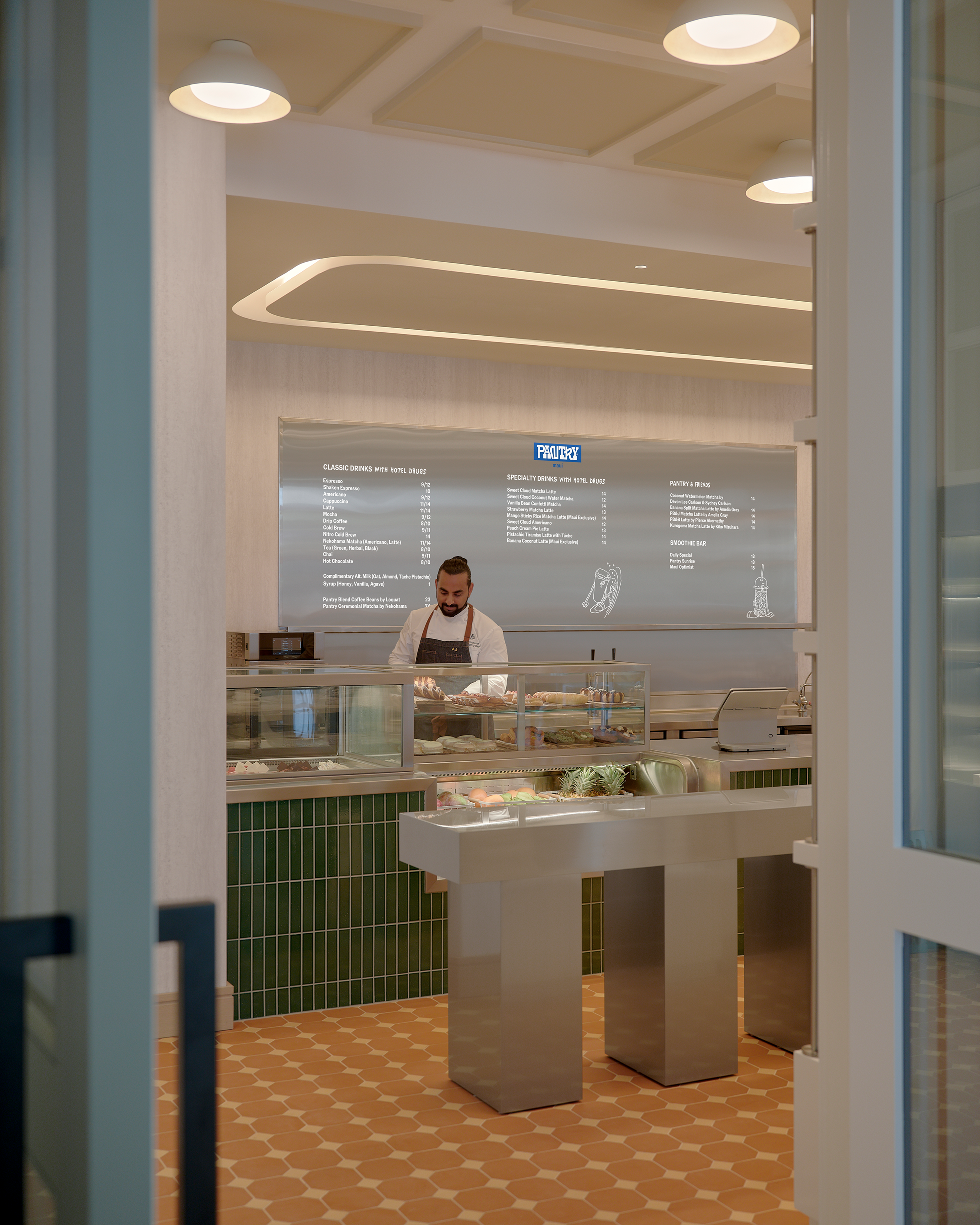 A man in an apron stands behind a modern deli counter with food displays, green tiled walls, and terracotta floor tiles, seen through an open doorway.